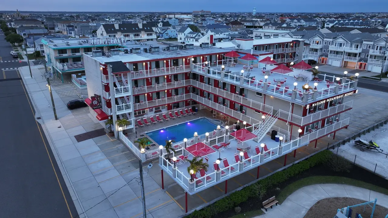 The Matador Oceanfront Resort at dusk, its lit pool and red-and-white balconies stacked four floors above the sundeck, with North Wildwood behind it